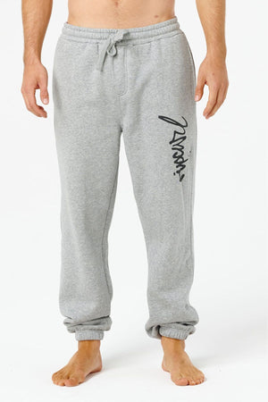 A person in light gray Rip Curl Icons Of Surf Trackpant sweatpants with a drawstring waist and black script on one leg stands barefoot against a plain white background.