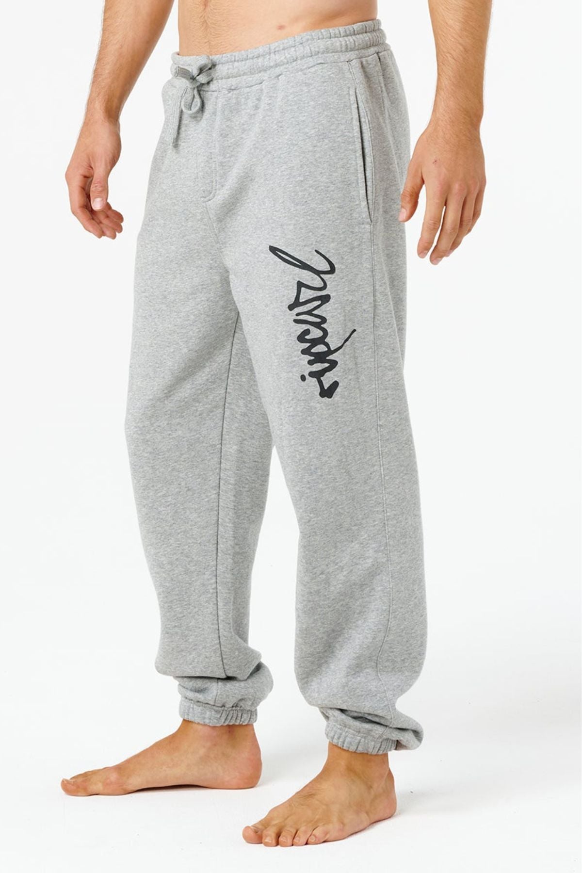 A person wearing Rip Curl Icons Of Surf Trackpant in light gray with an elastic waistband, ankle cuffs, side pockets, and black cursive text on the left leg stands barefoot on a white background.