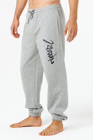 A person wearing Rip Curl Icons Of Surf Trackpant in light gray with an elastic waistband, ankle cuffs, side pockets, and black cursive text on the left leg stands barefoot on a white background.