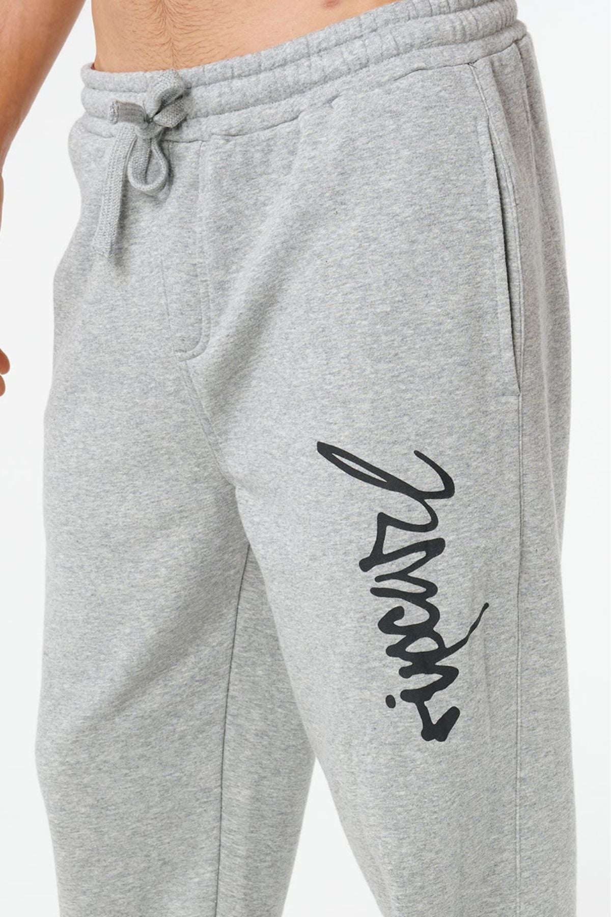 A person wearing Rip Curl Icons Of Surf Trackpant in light grey, featuring a drawstring waist, side pockets, and black cursive writing on the left thigh. Only their lower torso and upper legs are visible.