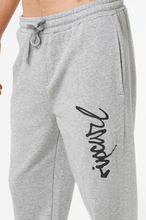 A person wearing Rip Curl Icons Of Surf Trackpant in light grey, featuring a drawstring waist, side pockets, and black cursive writing on the left thigh. Only their lower torso and upper legs are visible.