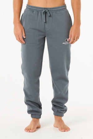 A person stands barefoot in blue-gray Rip Curl Icons Of Surf Trackpant sweatpants featuring a drawstring waist, side pockets, straight fit, and a small white and red logo on the upper left thigh.