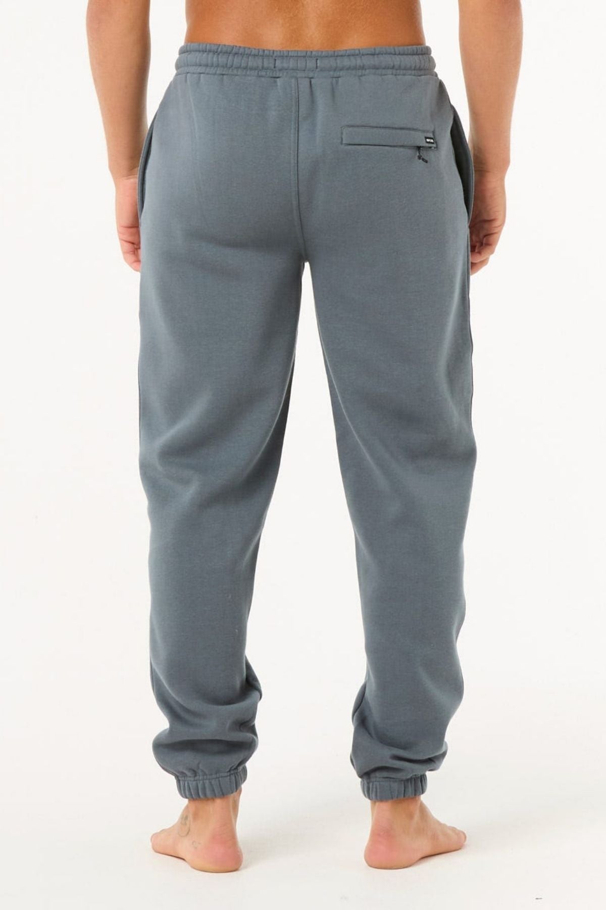 A person stands barefoot, facing away from the camera, wearing light blue-gray Rip Curl Icons Of Surf Trackpant joggers featuring an elastic waistband, side pockets, zippered back pocket, and elastic ankle cuffs.