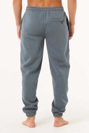 A person stands barefoot, facing away from the camera, wearing light blue-gray Rip Curl Icons Of Surf Trackpant joggers featuring an elastic waistband, side pockets, zippered back pocket, and elastic ankle cuffs.