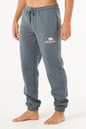 A person stands barefoot wearing Rip Curl Icons Of Surf Trackpant joggers with a straight fit, elastic waistband and cuffs, side pockets, and a white and pink "Rip Curl" logo on the left thigh. Upper body not visible.