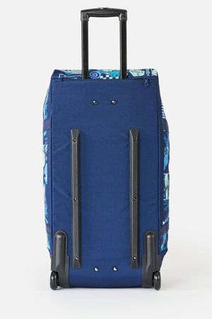 The Rip Curl Jupiter 80L Mixed Travel Bag stands upright with an extended black handle, black plastic rails, and two wheels. It has patterned blue accents on the sides and top, and is made from recycled polyester.
