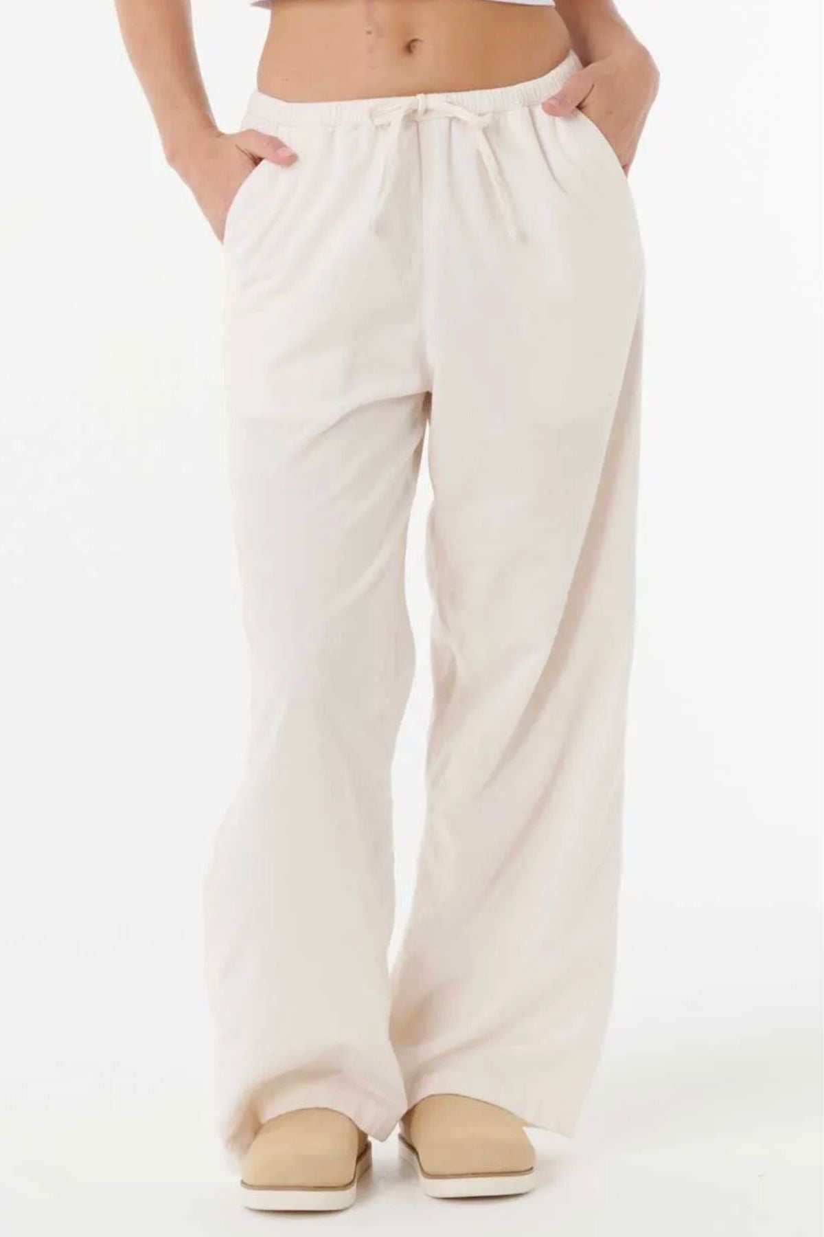 Someone stands against a white background wearing the Rip Curl Keepsakes Washed Cord Pant, a loose-fitting, light beige cotton corduroy pant with an elastic waistband and hands in pockets. The upper body is mostly out of frame. Beige shoes complete the look.