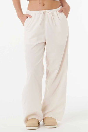 Someone stands against a white background wearing the Rip Curl Keepsakes Washed Cord Pant, a loose-fitting, light beige cotton corduroy pant with an elastic waistband and hands in pockets. The upper body is mostly out of frame. Beige shoes complete the look.