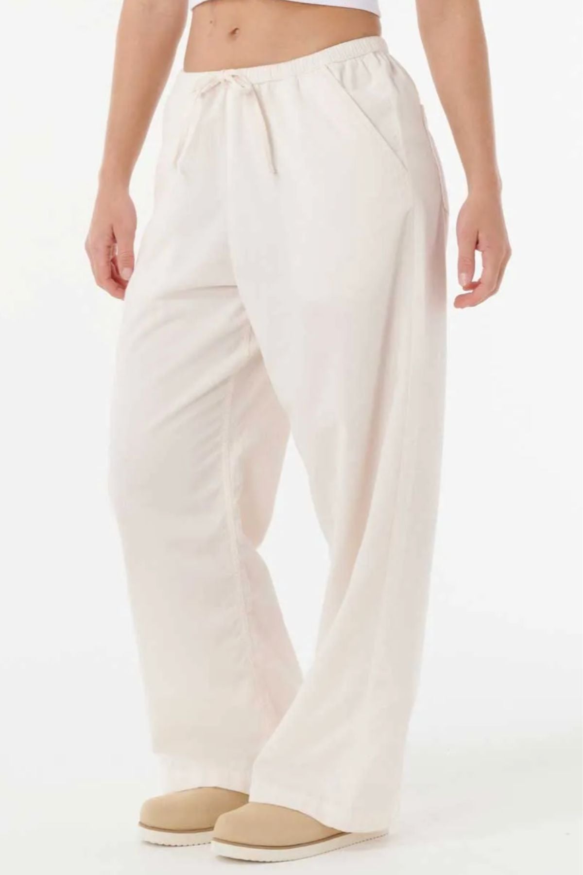 A person wearing the Rip Curl Keepsakes Washed Cord Pant—loose-fitting, high-waisted, off-white cotton with an elastic waistband and side pockets—pairs it with beige slip-on shoes and stands against a white background.