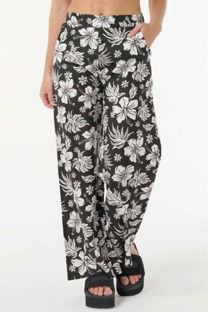A person wearing Rip Curl Keepsakes Beach Pant—high-waisted, wide-leg, black with a white tropical floral print—paired with black platform sandals, poses with one hand in the pocket against a plain white background.