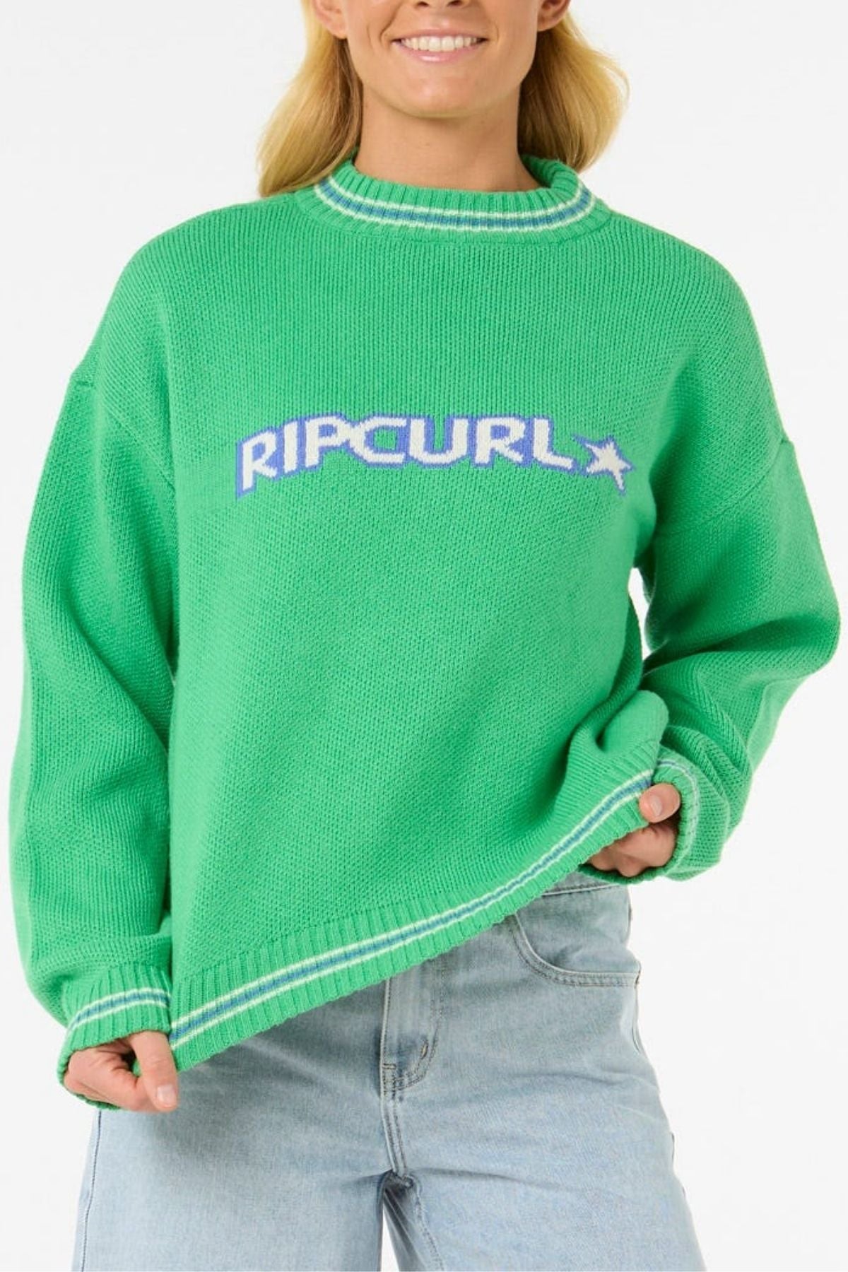 Wearing the cozy and stylish Rip Curl Las Dalias Sweater in bright green with light blue and white lettering, paired with light blue jeans, a person smiles in front of a plain white background.