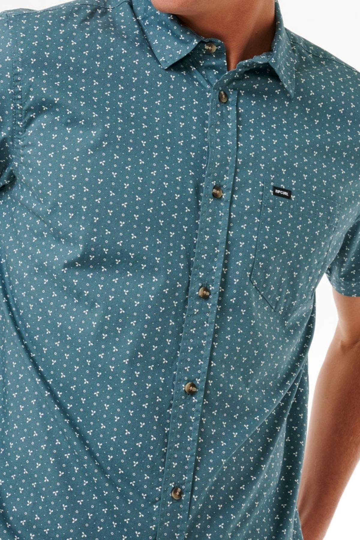 A person wears the Rip Curl Micro Short Sleeve Shirt in teal, featuring a small white floral print, chest pocket with a black label, brown buttons, and cotton blend fabric. The person's face is not shown.