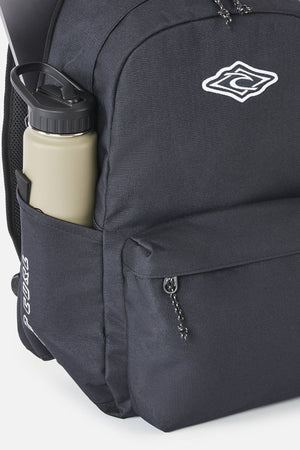 Black Rip Curl Crusher 30L Classic Backpack with a front zip pocket, a side water bottle sleeve containing a beige bottle, and a white graphic logo on the upper front.