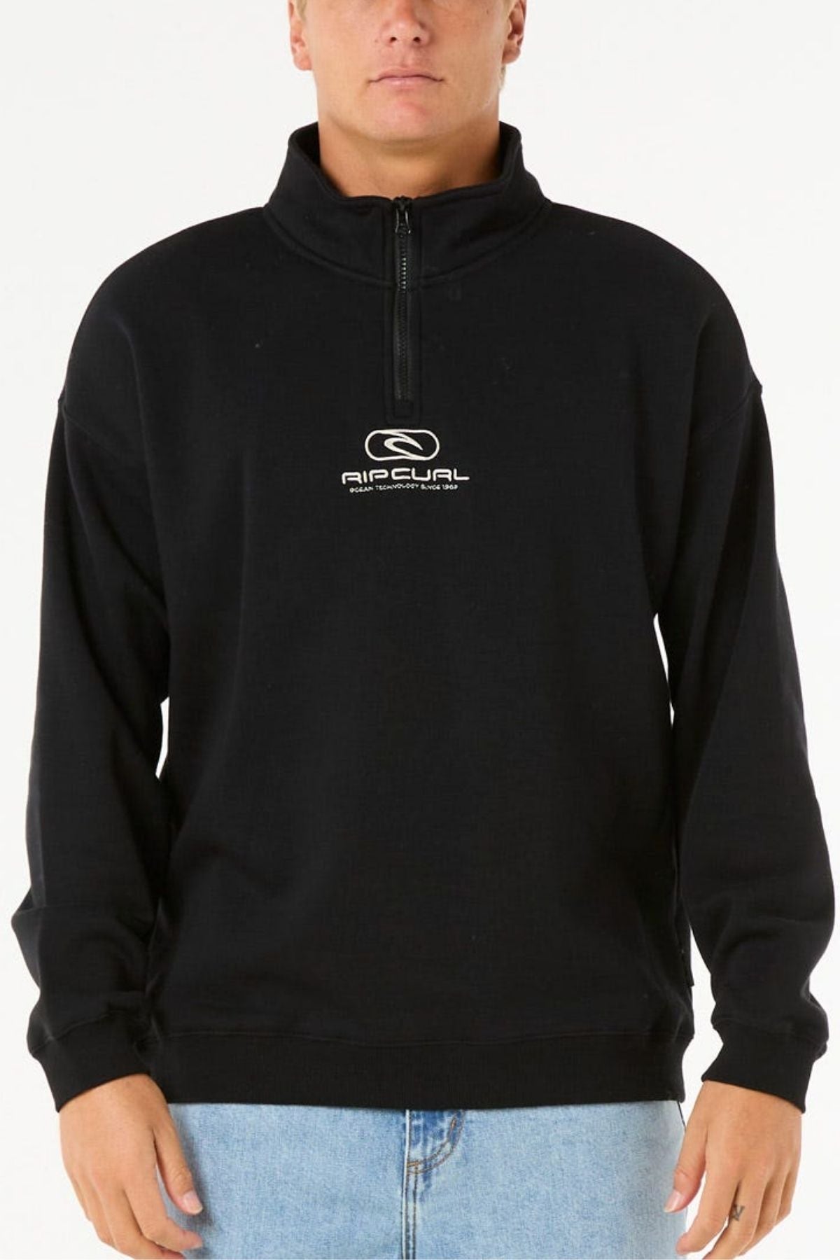 A person stands against a plain white background wearing the Rip Curl Pill Icon 1/4 Zip Crew, a black pullover made from Organic Cotton Poly Fleece with chest embroidery, paired with light blue jeans.