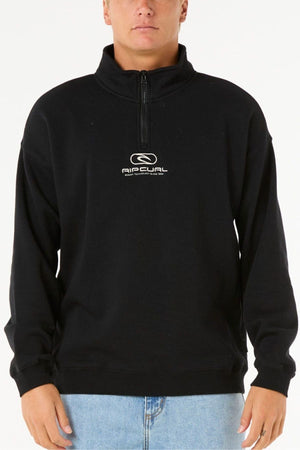 A person stands against a plain white background wearing the Rip Curl Pill Icon 1/4 Zip Crew, a black pullover made from Organic Cotton Poly Fleece with chest embroidery, paired with light blue jeans.