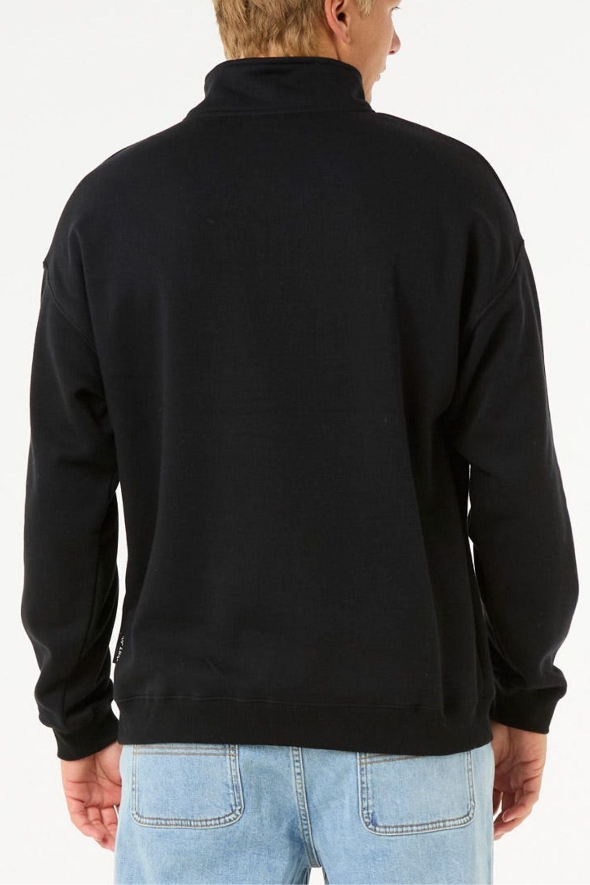 A person with short blonde hair is seen from the back, wearing a black Rip Curl Pill Icon 1/4 Zip Crew sweatshirt with chest embroidery and light blue jeans against a plain white background.