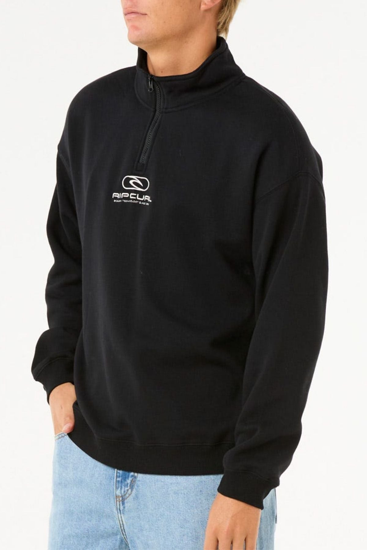 A person wearing a black Rip Curl Pill Icon 1/4 Zip Crew with chest embroidery and light blue jeans stands with one hand in their pocket against a plain white background.