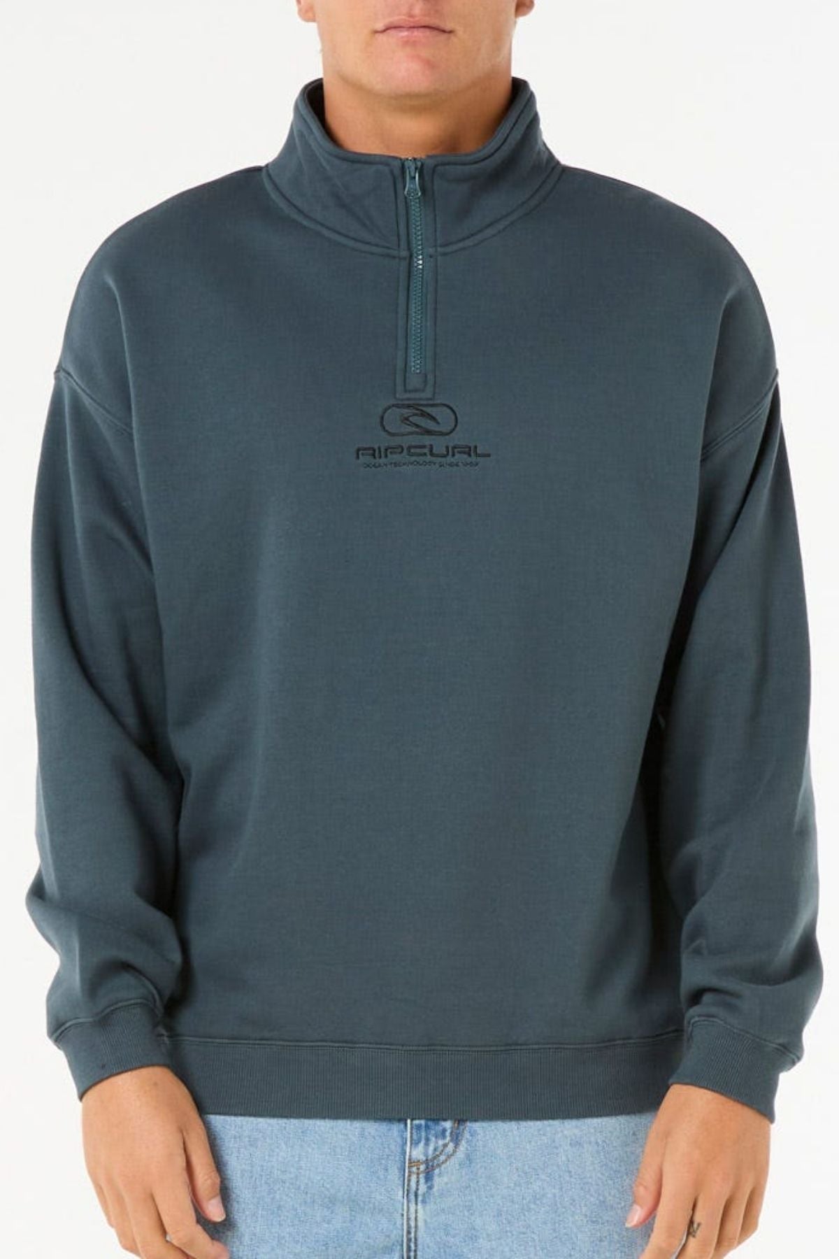 A person wearing a Rip Curl Pill Icon 1/4 Zip Crew in dark teal with "RIP CURL" and a logo on the chest, paired with light blue jeans, stands against a plain white background.