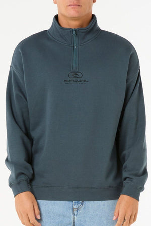 A person wearing a Rip Curl Pill Icon 1/4 Zip Crew in dark teal with "RIP CURL" and a logo on the chest, paired with light blue jeans, stands against a plain white background.