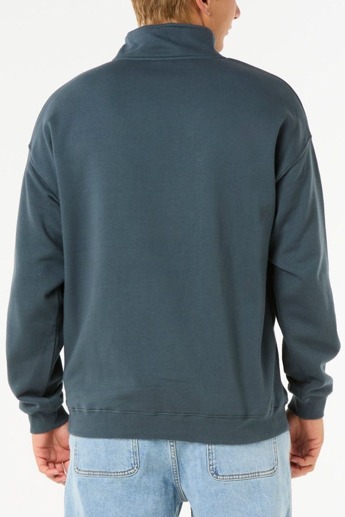 A person is seen from behind, wearing a Rip Curl Pill Icon 1/4 Zip Crew sweatshirt in dark teal and light blue jeans against a plain white background.