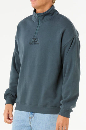 A person is wearing a dark teal Rip Curl Pill Icon 1/4 Zip Crew sweatshirt and light blue jeans, standing with arms relaxed by their sides against a plain white background.