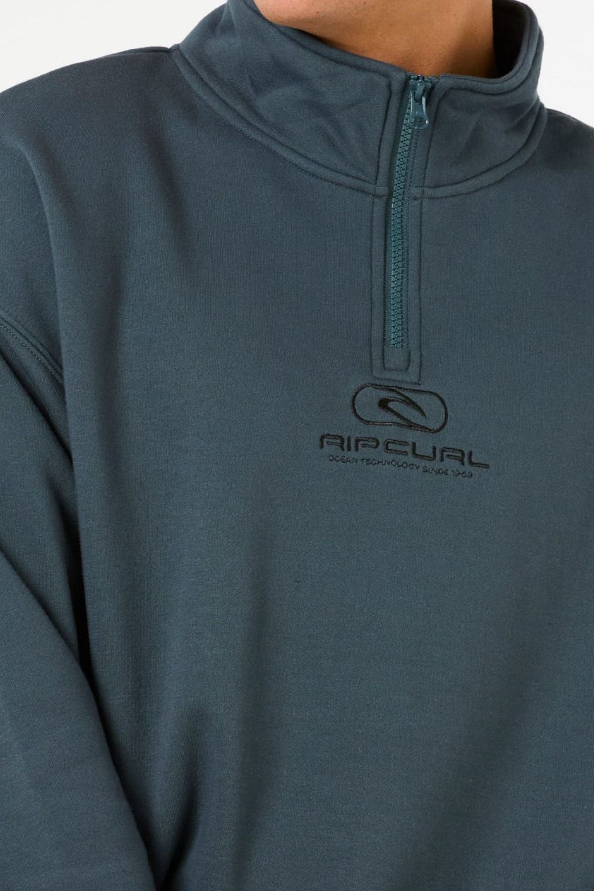 Someone wearing a teal Rip Curl Pill Icon 1/4 Zip Crew pullover with a high collar, made from Organic Cotton Poly Fleece. The chest displays the Rip Curl logo and "Ocean Technology Since 1969." The person’s face is mostly out of frame.