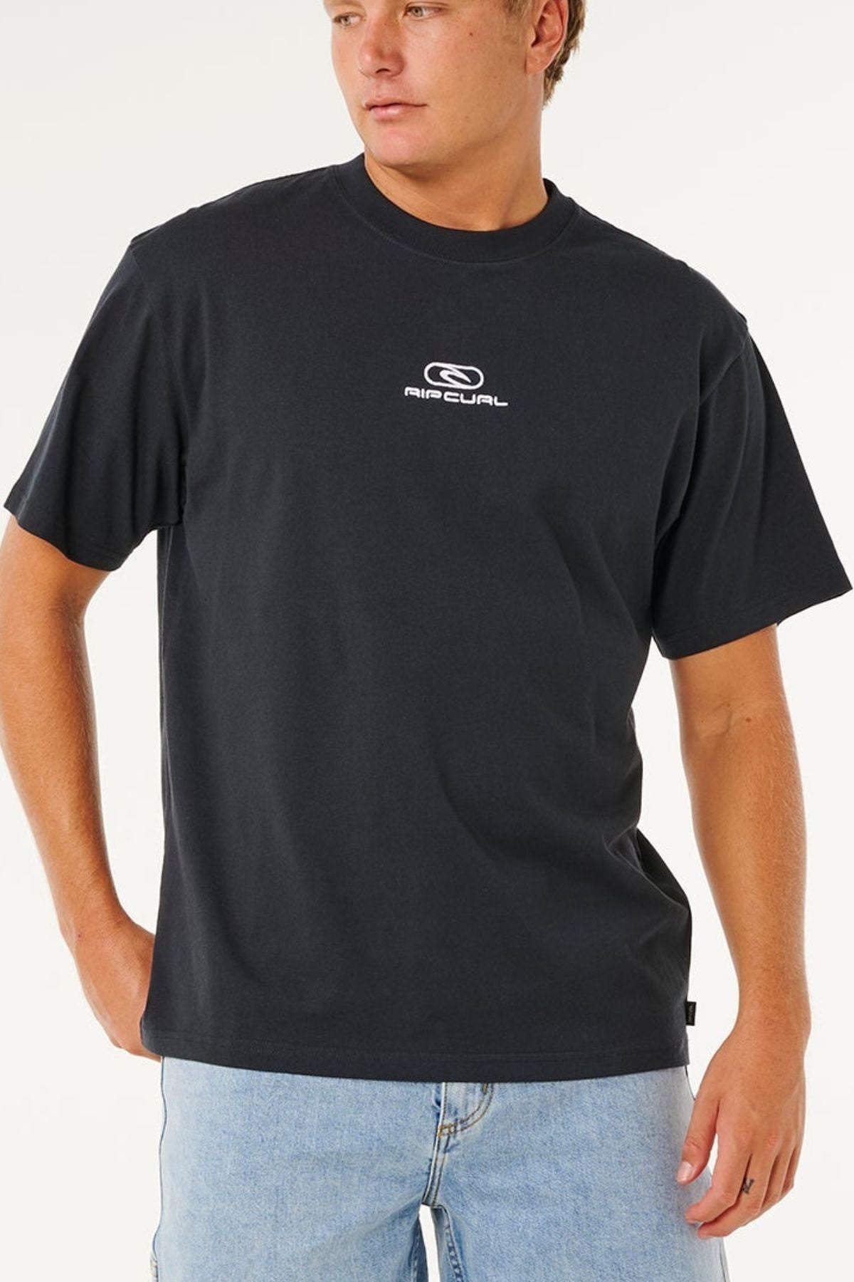 Wearing a relaxed, loose fit Rip Curl Pill Icon Tee in black made from responsibly sourced cotton and light blue jeans, a person stands with one hand in their pocket, gazing to the side against a plain background.