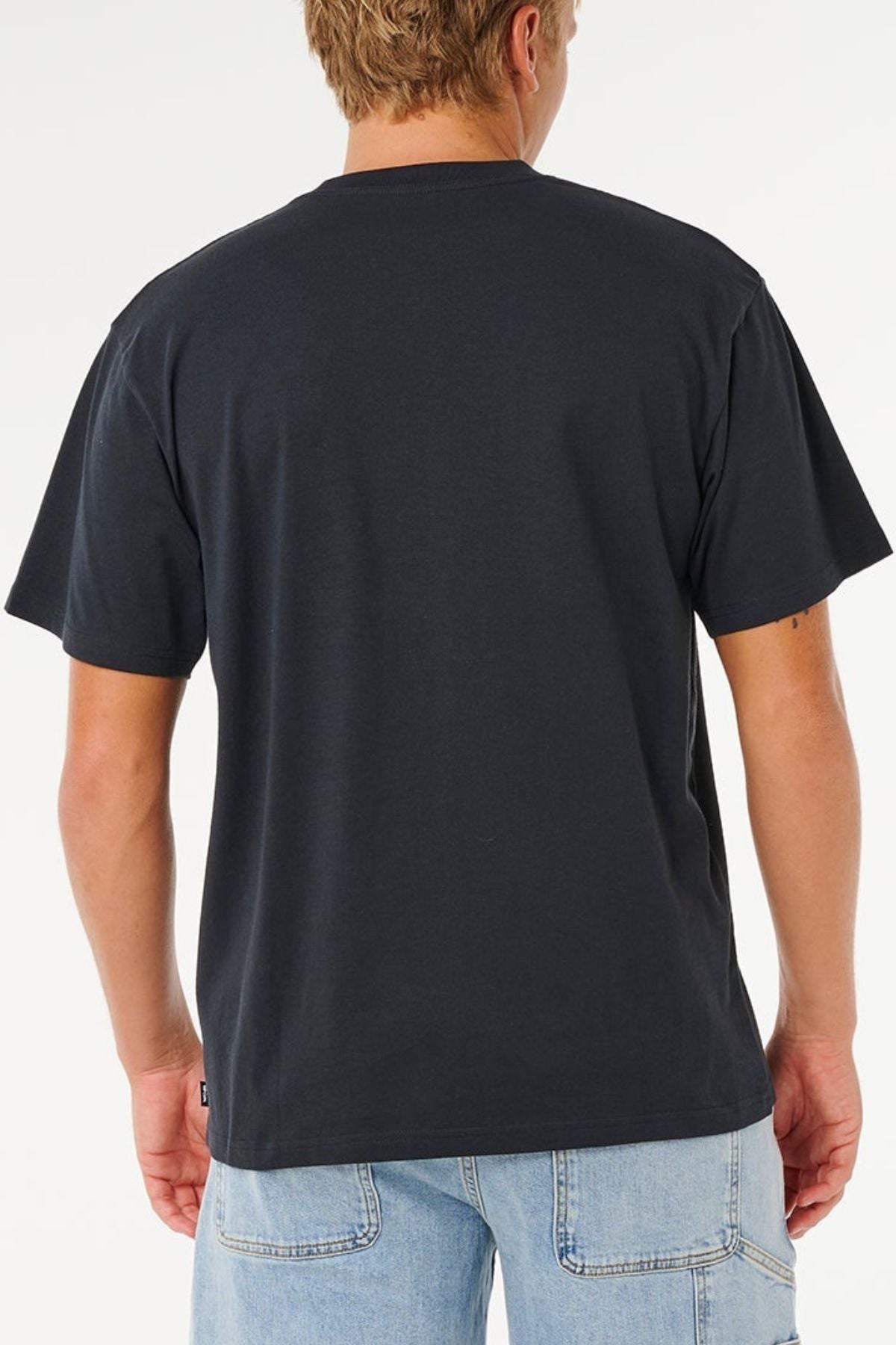 A person with short blonde hair, dressed in a relaxed loose fit plain black Rip Curl Pill Icon Tee made from responsibly sourced cotton and light blue jeans, stands facing away from the camera against a white background.