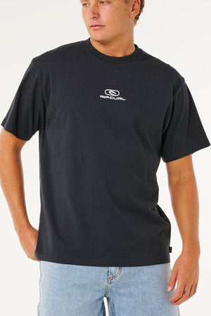 Wearing a relaxed, loose fit Rip Curl Pill Icon Tee in black made from responsibly sourced cotton and light blue jeans, a person stands with one hand in their pocket, gazing to the side against a plain background.