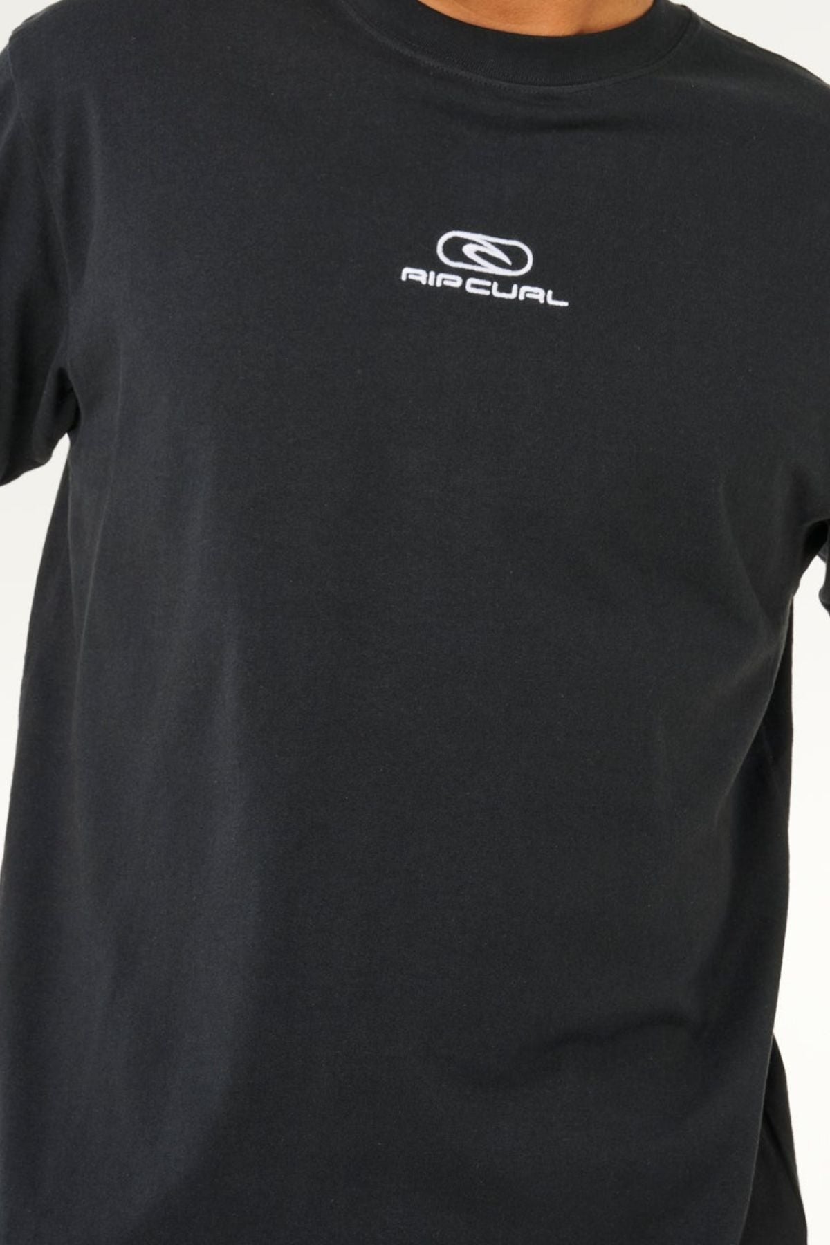 A person is wearing the Rip Curl Pill Icon Tee, a plain black t-shirt featuring a small white Rip Curl logo and icon design on the chest. Made from responsibly sourced cotton and designed with a relaxed, loose fit, the tee is shown from shoulders to waist against a simple background.
