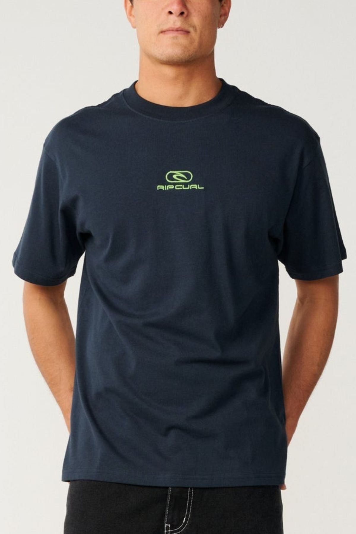 A person stands against a plain, light background wearing the Rip Curl Pill Icon Tee in dark blue, which features a small neon green Rip Curl logo and text on the chest. The tee is crafted from responsibly sourced cotton and designed with a relaxed loose fit.