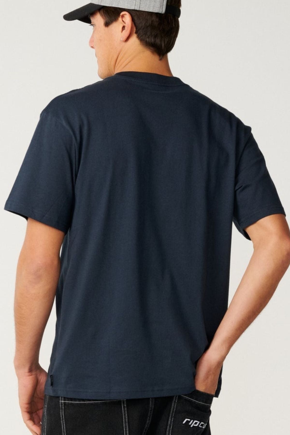 Wearing the Rip Curl Pill Icon Tee, a man stands with his back to the camera, one hand partially in his pocket. The relaxed, loose fit and responsibly sourced cotton of this t-shirt add effortless style to his laid-back look.