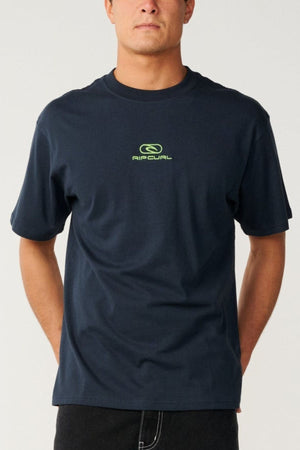 A person stands against a plain, light background wearing the Rip Curl Pill Icon Tee in dark blue, which features a small neon green Rip Curl logo and text on the chest. The tee is crafted from responsibly sourced cotton and designed with a relaxed loose fit.