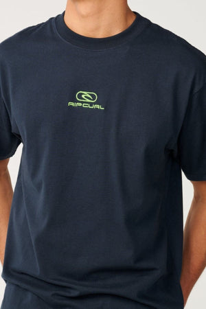 Wearing the Rip Curl Pill Icon Tee in navy blue, this individual sports a relaxed, loose-fitting top made from responsibly sourced cotton. The t-shirt features a small green logo with text on the chest. The person’s head is not visible in the image.