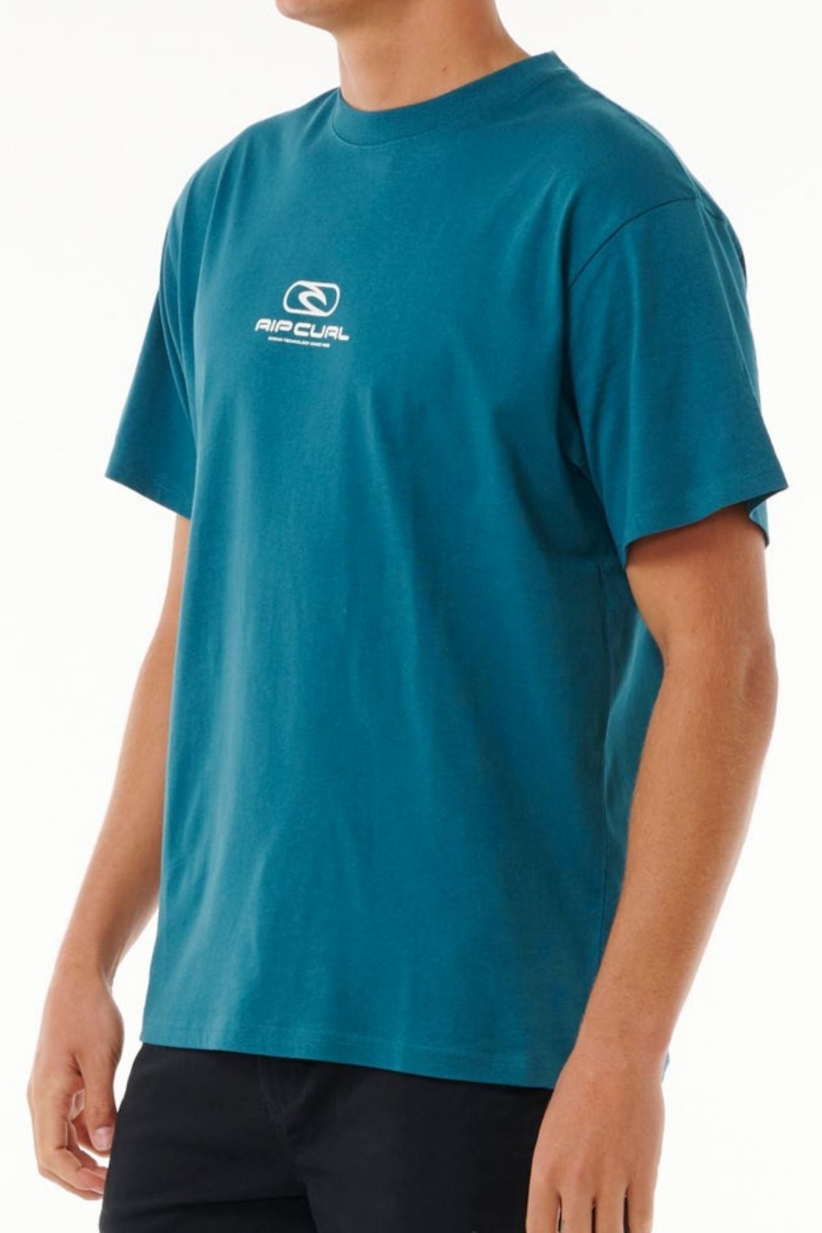 A person wears a teal Rip Curl Pill Icon Tee, 100% cotton, featuring the white logo and text on the chest, paired with black pants against a plain white background.