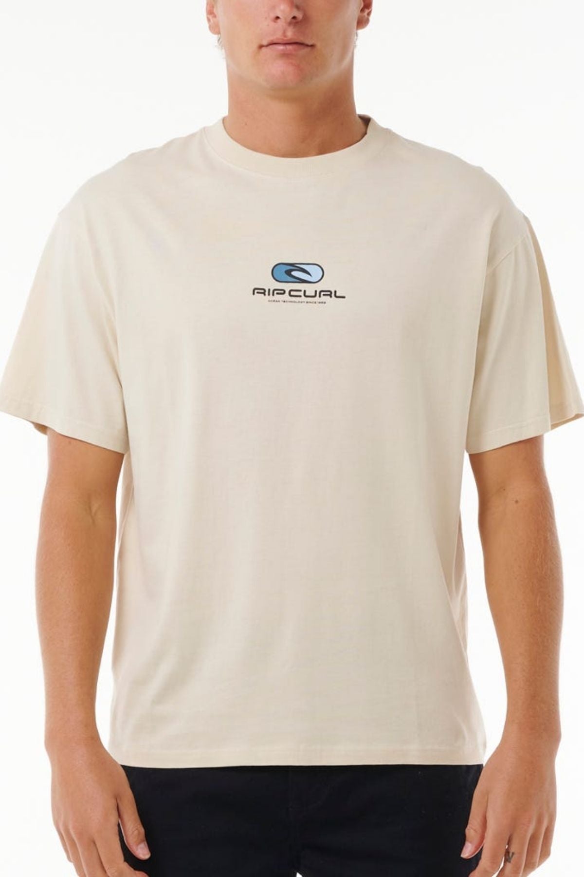 A person wears a beige "Rip Curl Pill Icon Tee" with a small centered "AIRUAL" logo and an abstract front design. It's classic and made of 100% cotton, set against a plain white background.