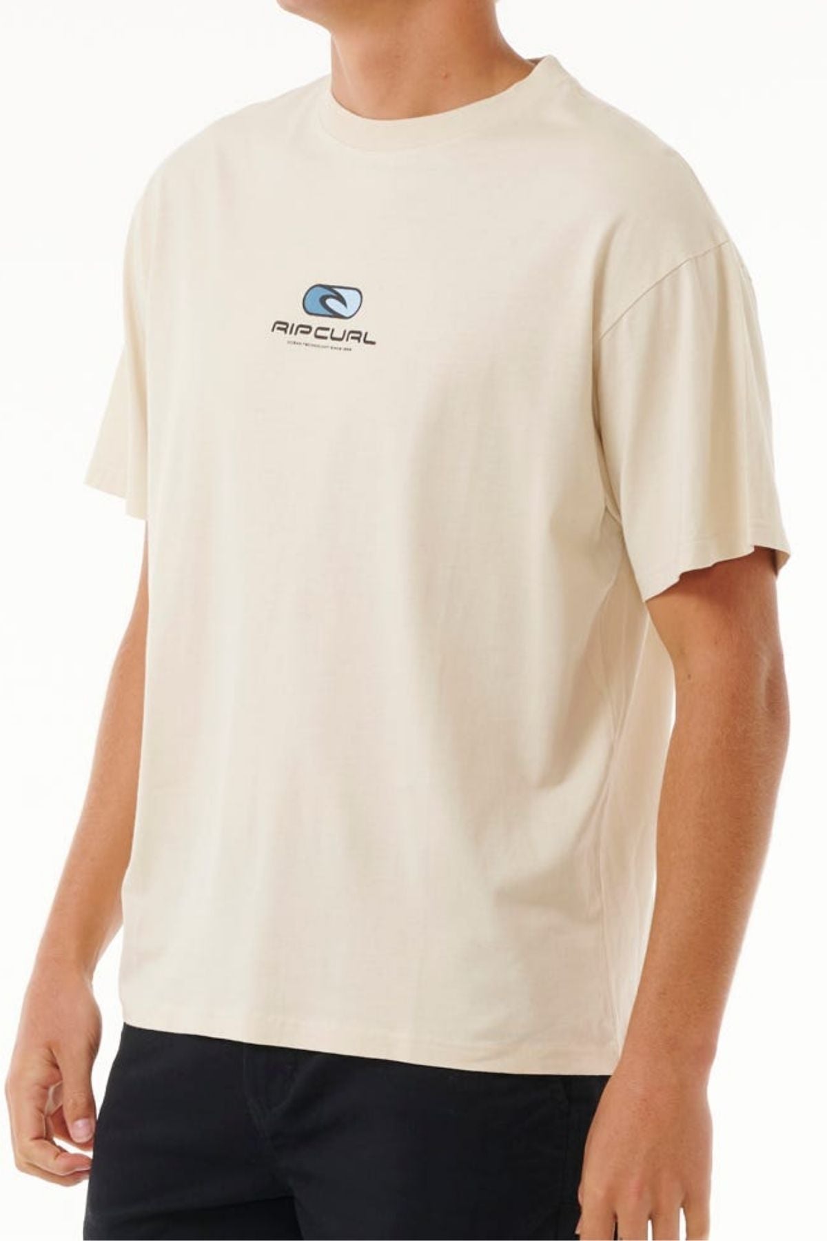 A person is wearing a Rip Curl Pill Icon Tee in beige, crafted from 100% cotton. It features a small chest logo and exudes classic style, effortlessly complementing dark pants against a plain, light background.
