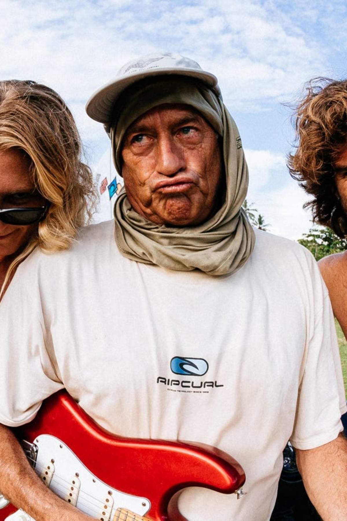 Three people huddle together under a blue sky with scattered clouds. The central figure wears a cap, scarf, and the Rip Curl Pill Icon Tee, making a funny face while holding a red electric guitar, adding to the classic style of the moment.