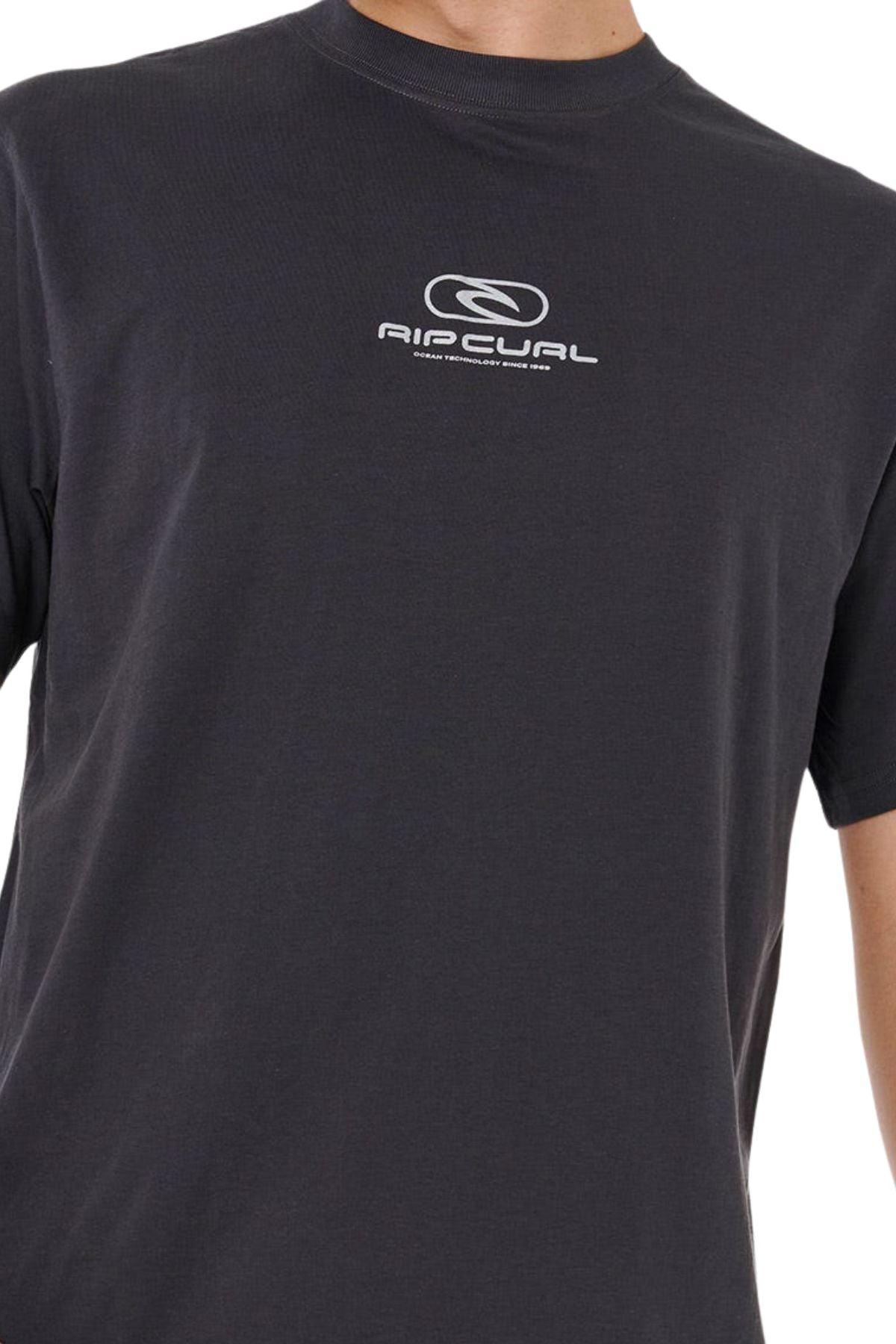 A person wears the Rip Curl Pill Icon Tee, a relaxed-fit dark gray shirt featuring a small white Rip Curl logo and text centered on the chest, shown from neck to waist with part of one arm visible.