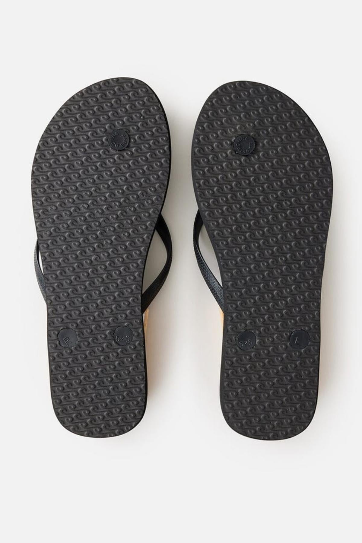 The image shows the bottom view of a pair of Rip Curl Premium Surf Bloom Thongs with textured soles. These black flip-flops feature simple thong-style straps against a plain white background.