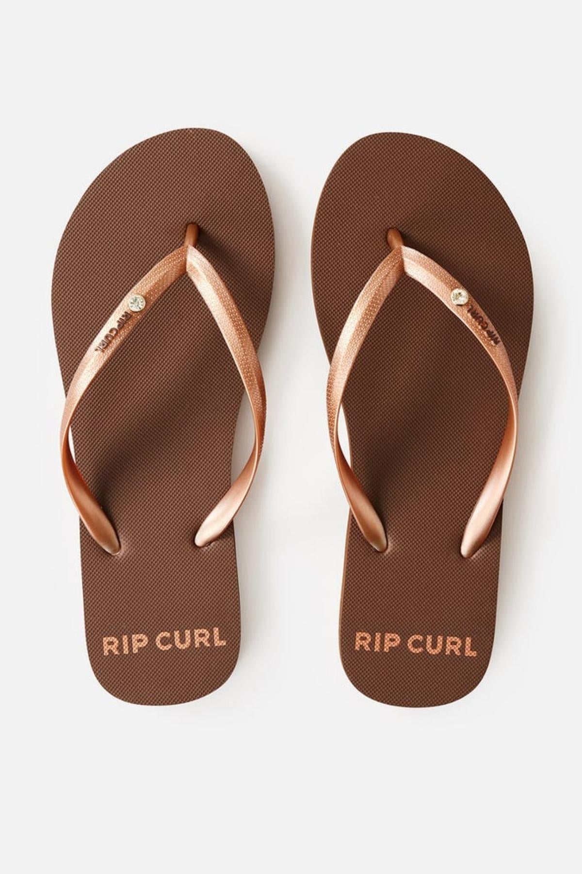 The Rip Curl Premium Surf Bloom Thongs feature a pair of brown flip-flops with gold straps. The text "RIP CURL" is elegantly printed in gold lettering on the footbed near the toe section of each sandal. Additionally, one of the straps on each sandal is adorned with a small circular metallic emblem. The background is white.