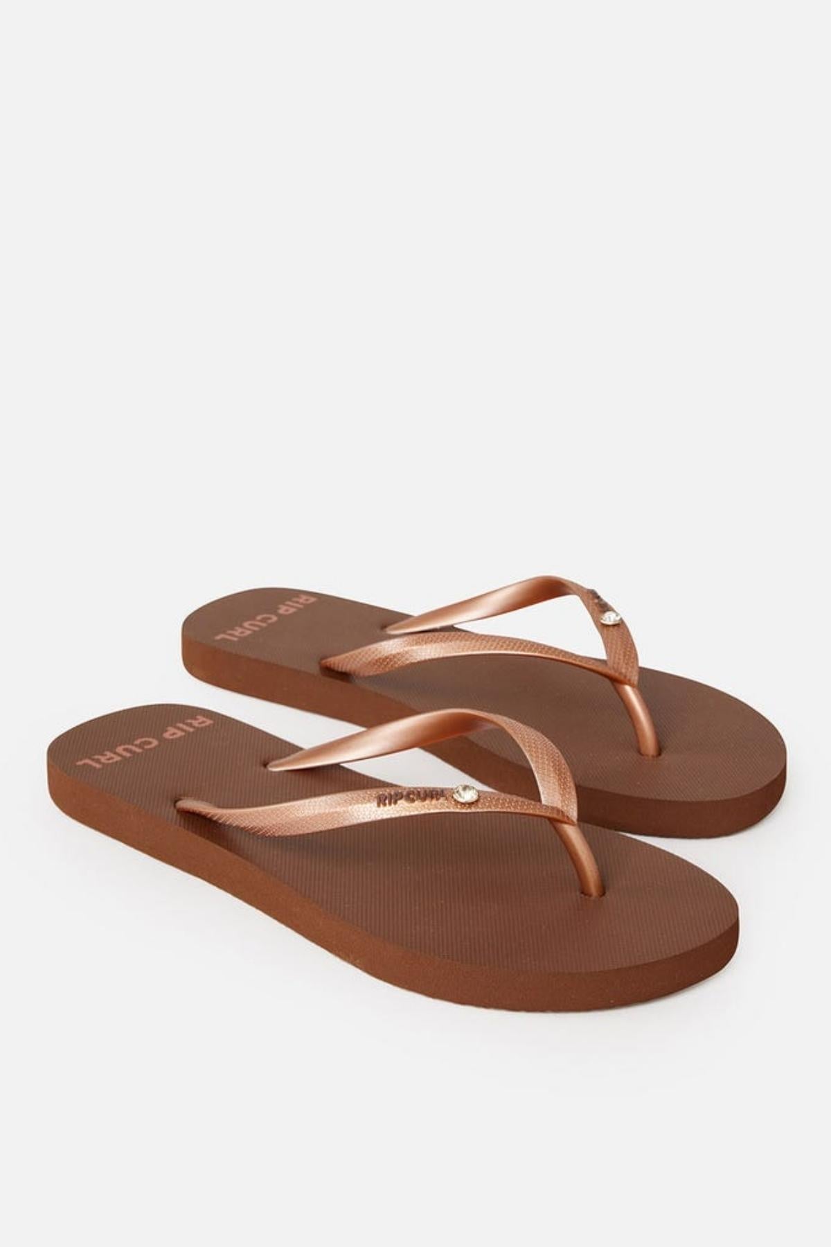 A pair of Rip Curl Premium Surf Bloom Thongs featuring bronze metallic straps with a small circular embellishment on each strap. The brown flip-flops have a textured sole, and the brand name "Rip Curl" is visible on the top of the sole near the heel. The background is plain white.