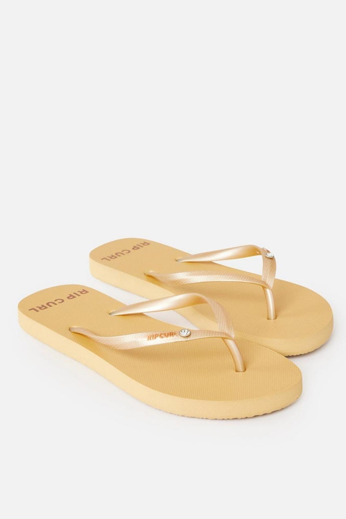 A pair of Rip Curl Premium Surf Bloom Thongs in beige with thin gold straps. The sole is slightly textured, and there is a small circular embellishment on each strap. The brand "Rip Curl" is visibly printed on the insole near the heel area. The background is plain white.