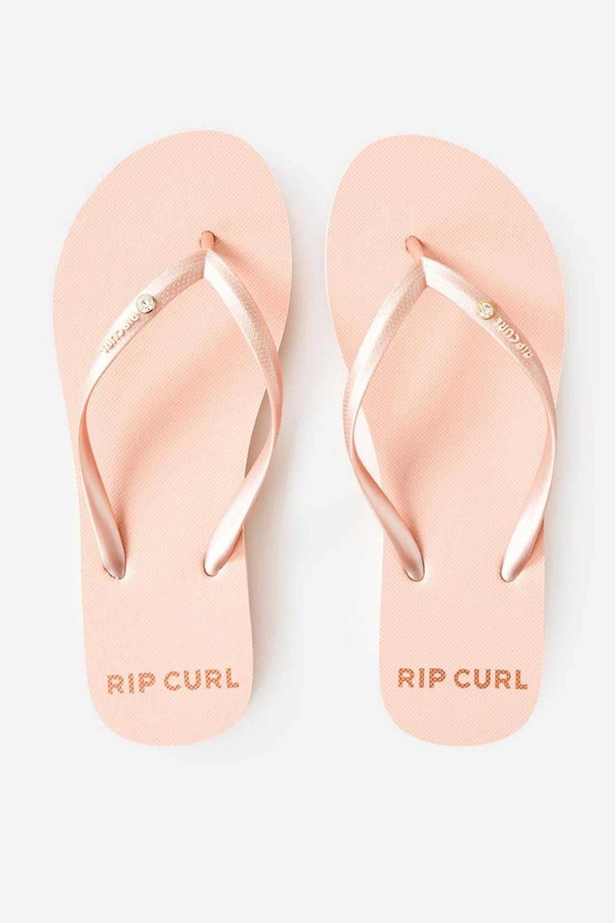 A pair of Rip Curl Premium Surf Bloom Thongs in light pink with matching metallic pink straps, featuring a small decorative button. The brand name "RIP CURL" is printed on the sole of each thong near the heel.