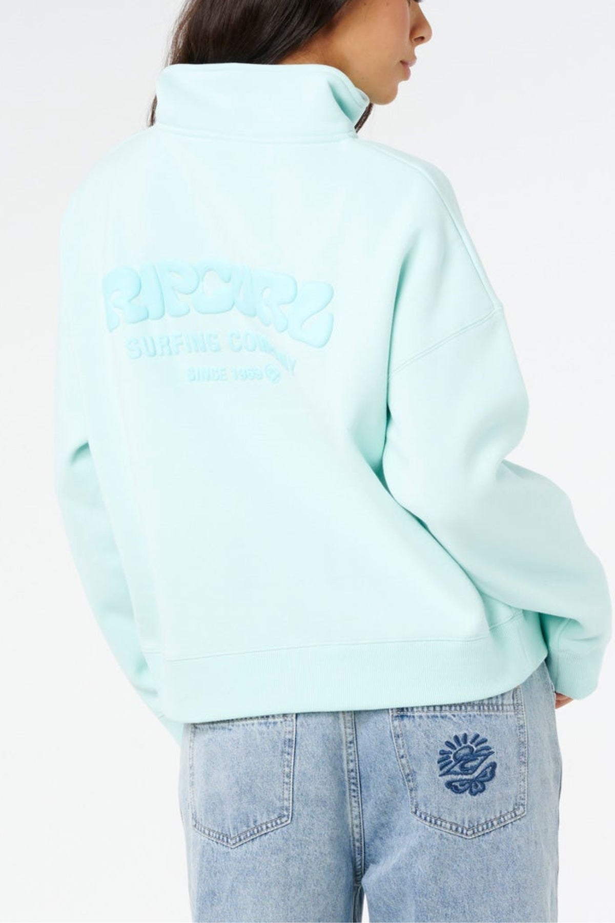 Wearing the Rip Curl Surf Puff 1/4 Zip in light blue with embossed back text and matching light blue jeans featuring a sun pocket design, this look channels surf style against a white background.