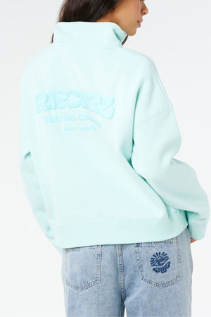 Wearing the Rip Curl Surf Puff 1/4 Zip in light blue with embossed back text and matching light blue jeans featuring a sun pocket design, this look channels surf style against a white background.