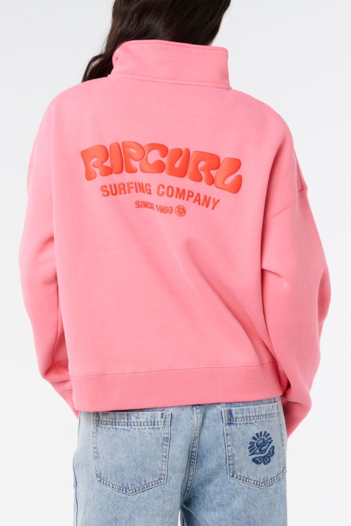 A person in a pink Rip Curl Surf Puff 1/4 Zip and light blue jeans, seen from the back against a white background, highlights relaxed surf-inspired style.