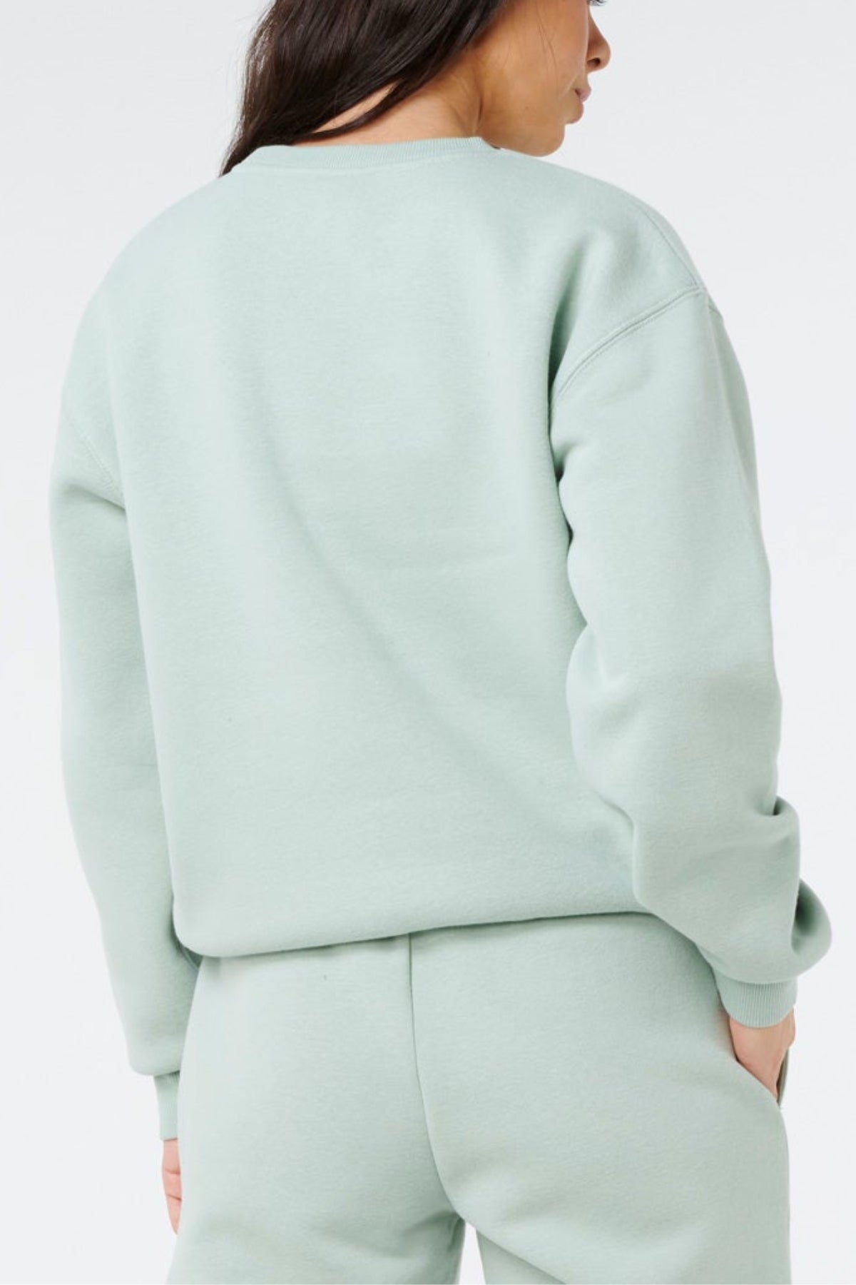 A woman is seen from the back, standing against a white background, wearing a light green Rip Curl Surf Staple Relaxed Crew sweatshirt and matching sweatpants.