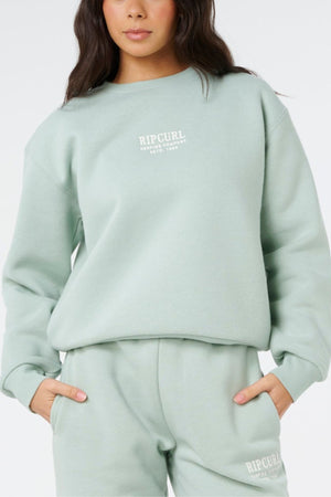 A person wears the Rip Curl Surf Staple Relaxed Crew and matching sweatpants in light green, featuring "Rip Curl" branding and a relaxed fit, standing with hands in pockets against a plain background.