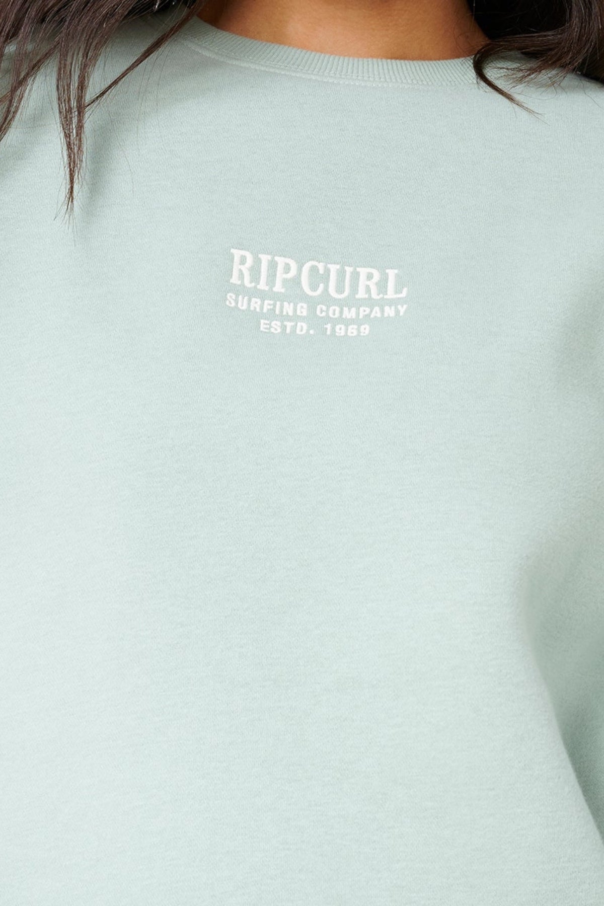 Close-up of a person in the Rip Curl Surf Staple Relaxed Crew pullover in light green with "RIP CURL SURFING COMPANY ESTD. 1969" in white on the chest, showing only the lower part of their face and body.